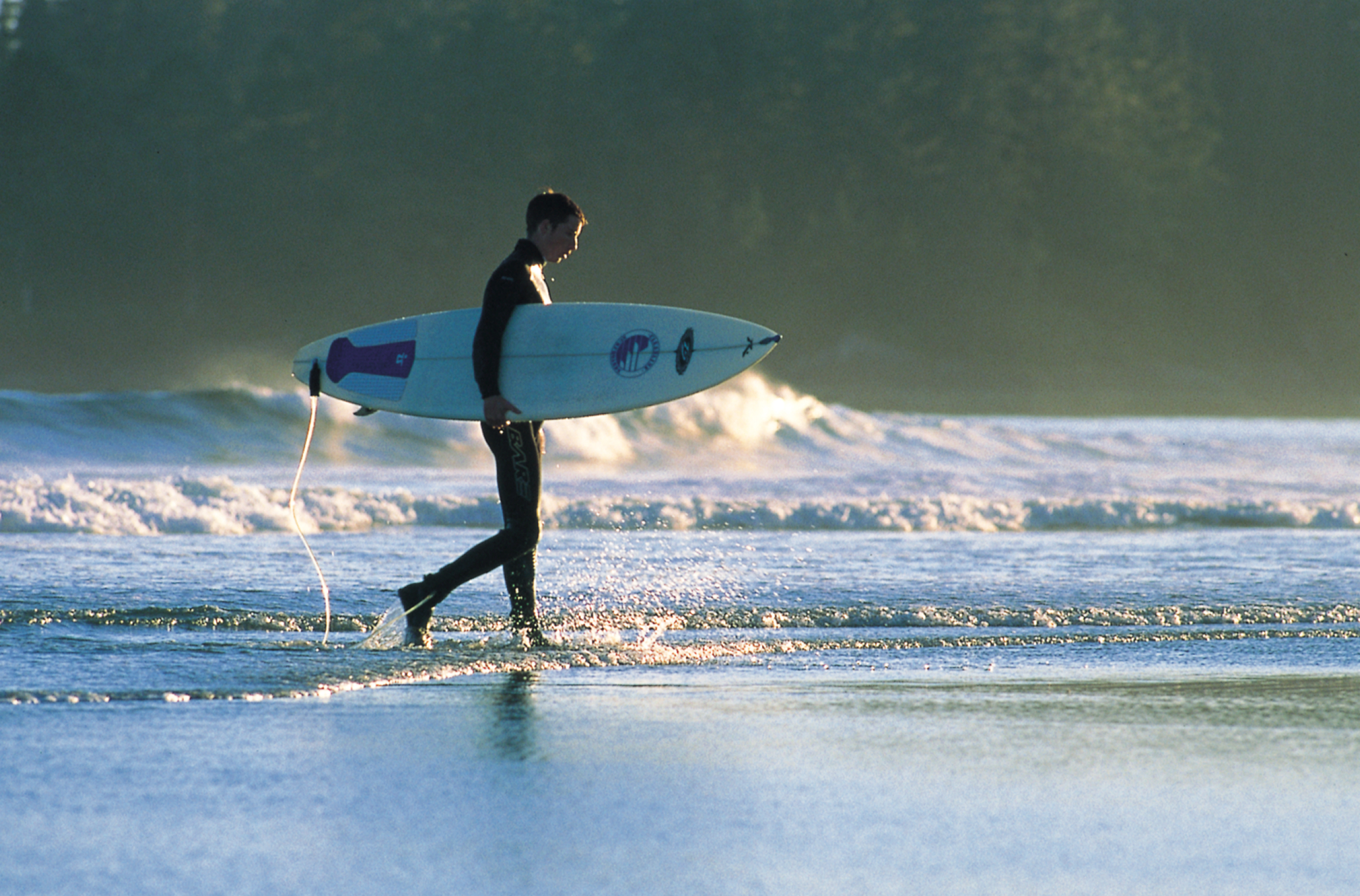 A man holds a surfboard under his arm as he walks along Long Beach in Tofino