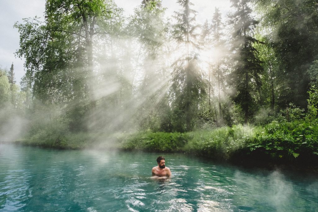 A man relaxes in a turquoise hot spring drenched in sunlight. filtering through the trees