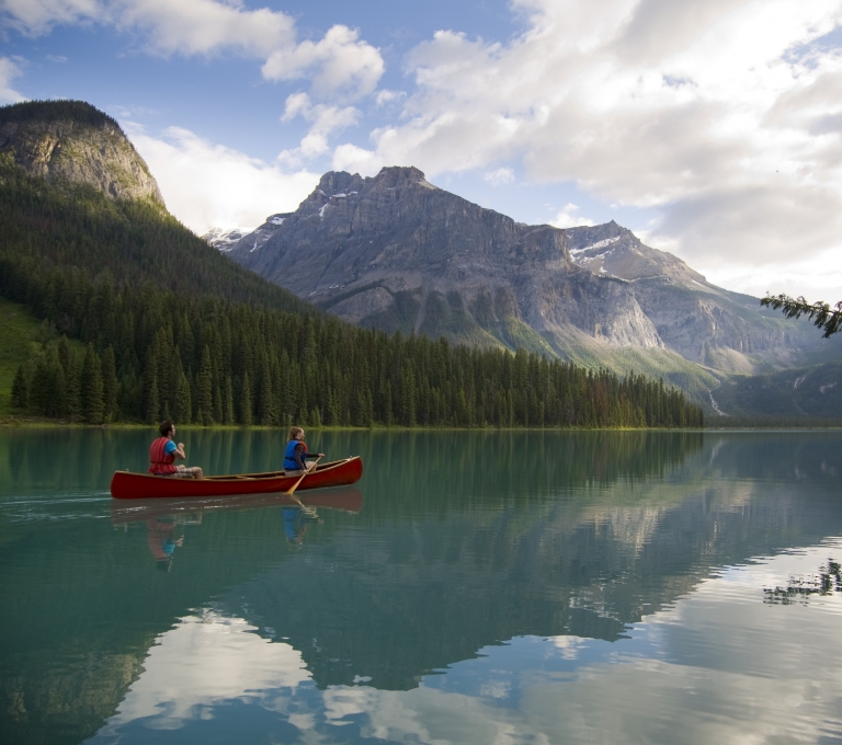 A red canoe is paddled by two people across a lake in Yoho National Park. The turquoise lake is surrounded by mountains and trees.