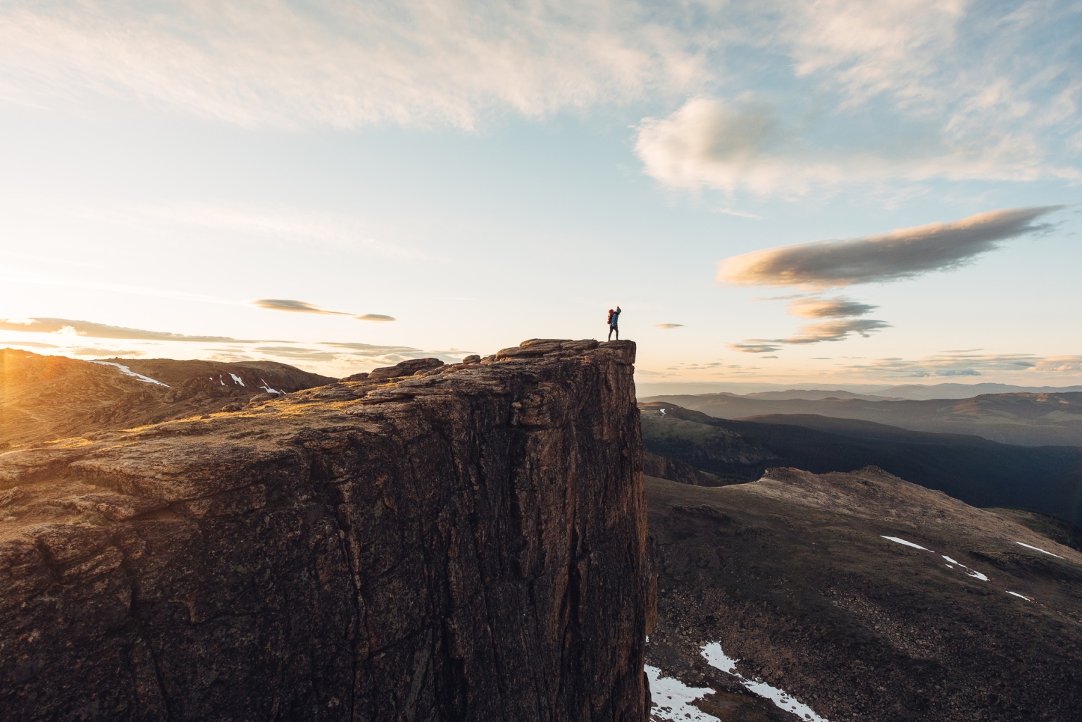 A person stands at the peak of a mountain as the sun rises behind them in Cathedral Provincial Park. They look out at the valley below.