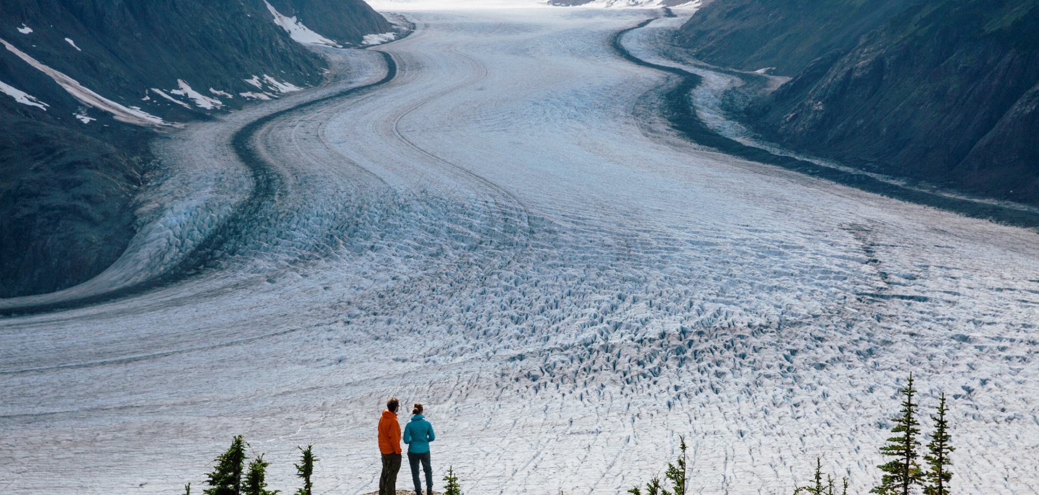 Two people look out at a frozen landscape in the Stewart-Cassiar region of northern BC. They stand on a rocky outcrop and are looking towards the mountain range.