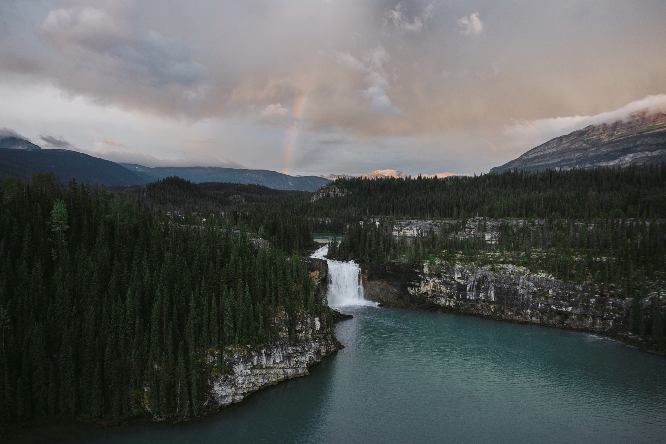 Waterfall between two mountain ranges with clouds overhead and a rainbow coming through the clouds in Northern BC