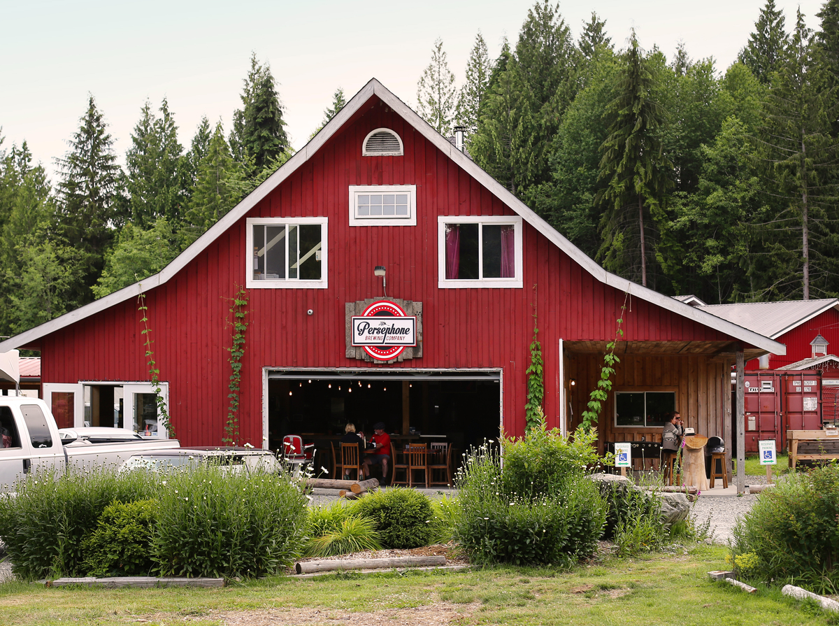 Outside the back of Persephone Brewing Company in Gibsons. The building is built like a large red barn with white trim. Tables are seen inside and cars and grass are out front.