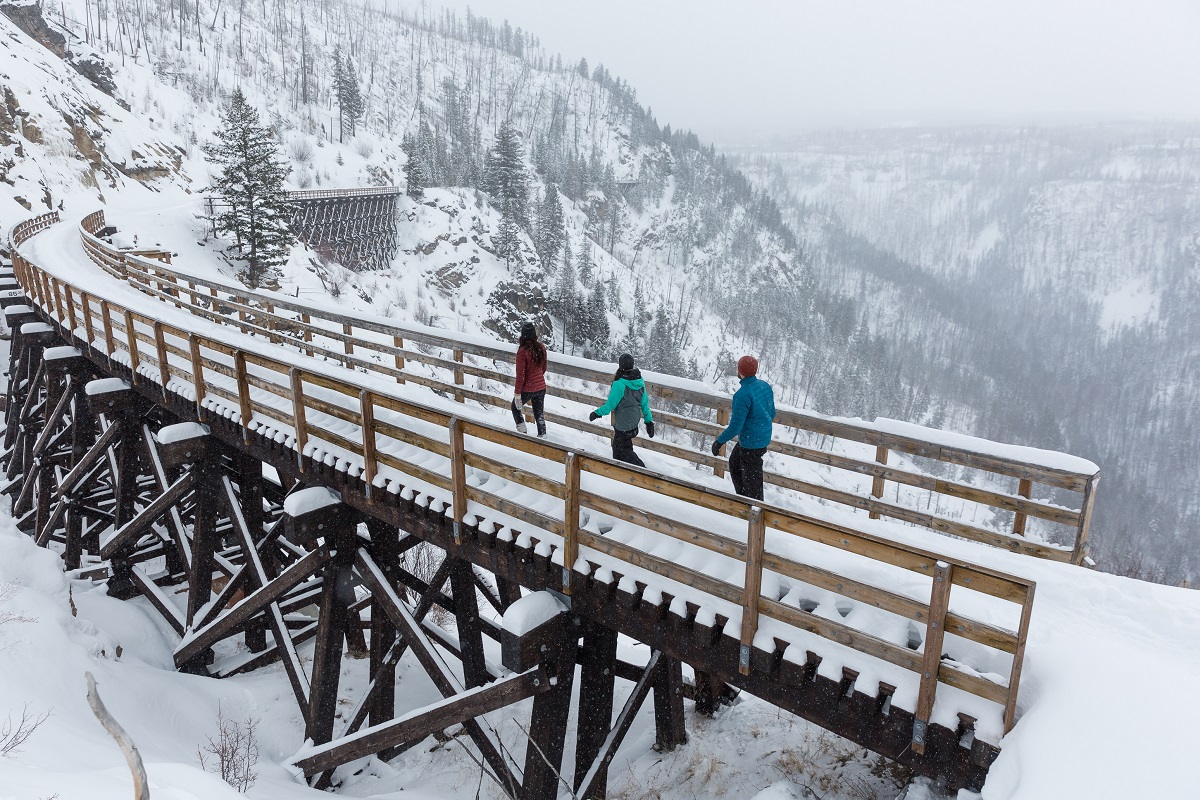 Three snowshoers walk across a wooden bridge covered in snow on the Kettle Valley Rail Trail