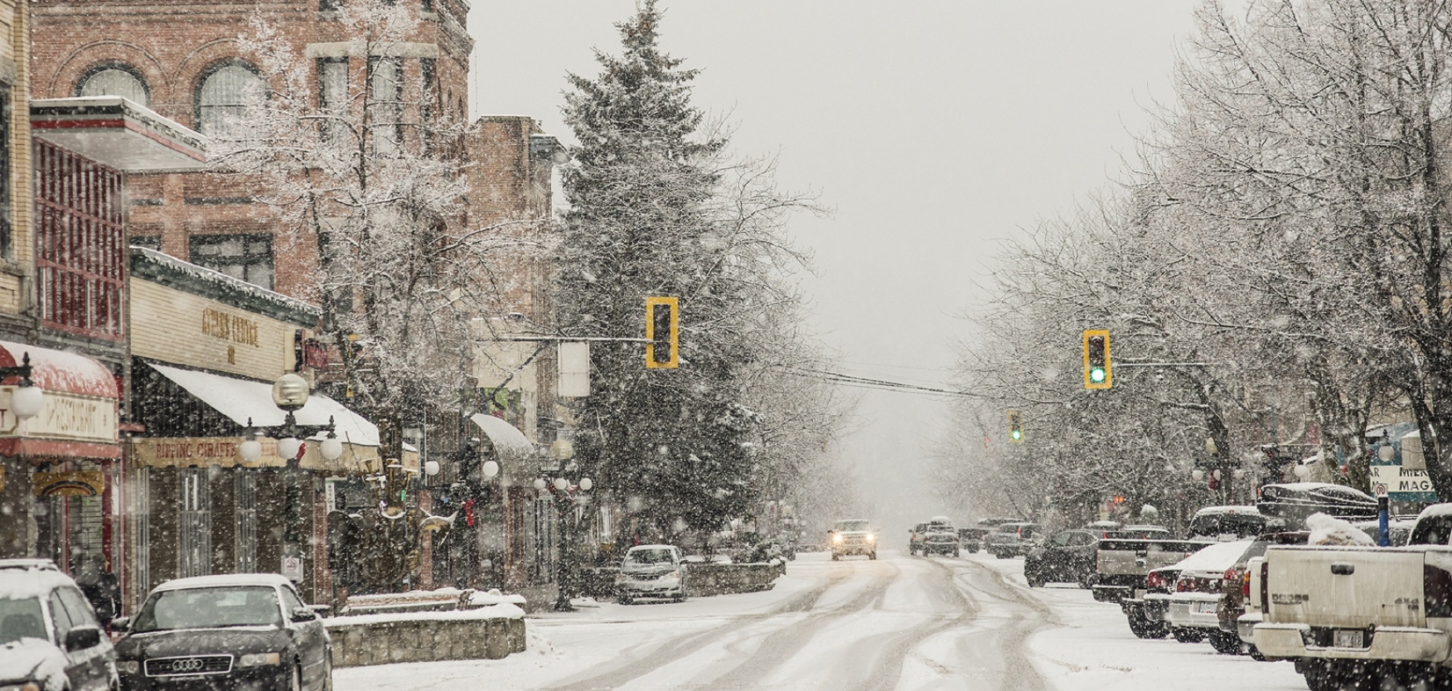 A snowy view of downtown Nelson. The snow covered street has cars on both sides. 