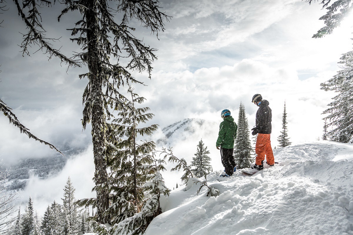 Two skiers stand on a snowy outlook looking down at the snowy run in front of them.