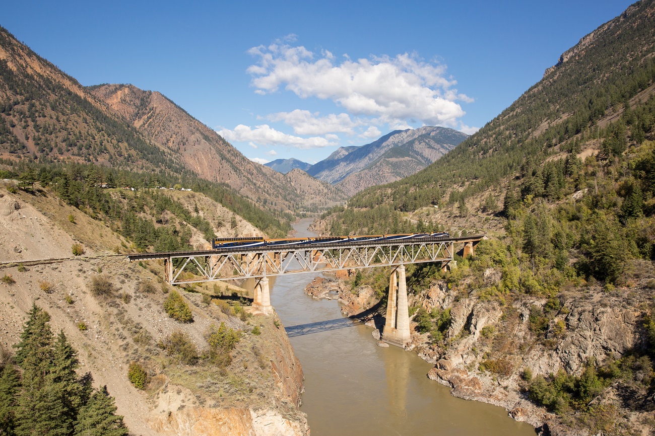 A train crosses a short bridge that crosses the brown, silty Fraser River. On the sides and in the distance, mountains with sparse vegetation.