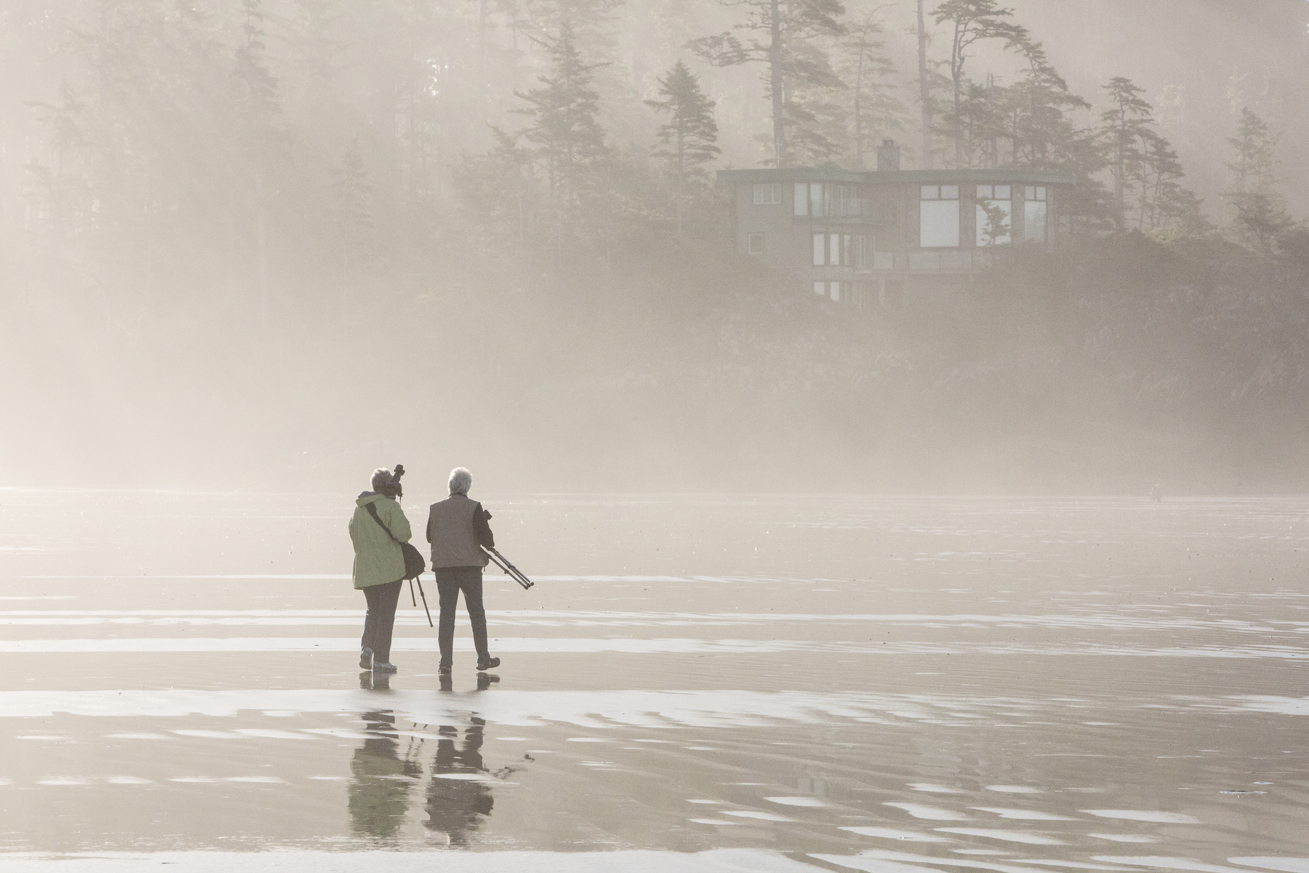 Two people walk along the beach at low tide in the fog. A building and trees are seen in the background.