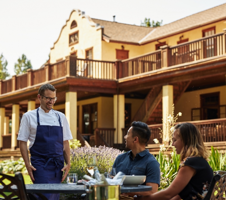 Chef Ned Bell describes the meal to a couple dining on the patio at the Naramata Inn. The building is two stories with a wrap around patio and gardens on the ground level.