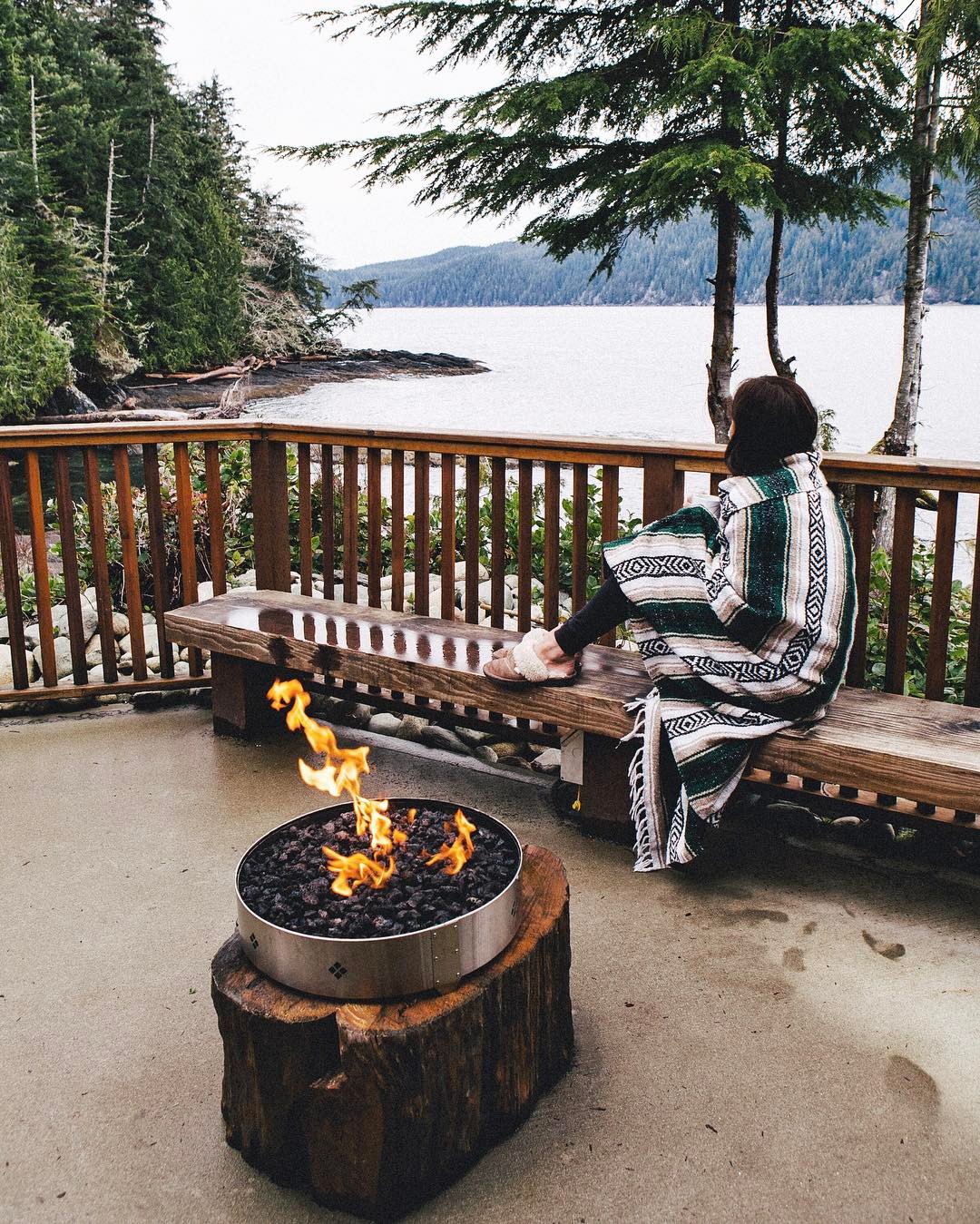 A woman is wrapped in a blanket, sitting on a patio beside a gas fire, overlooking the ocean behind her. There are trees on the shoreline.
