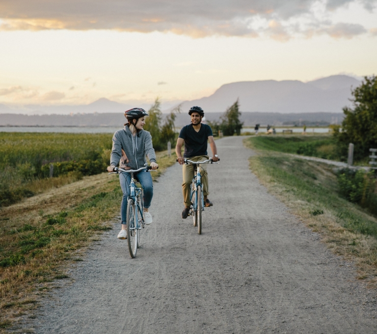 Two people ride bicycles down a flat gravel path. Fields and farmland are on both sides of the pathway.