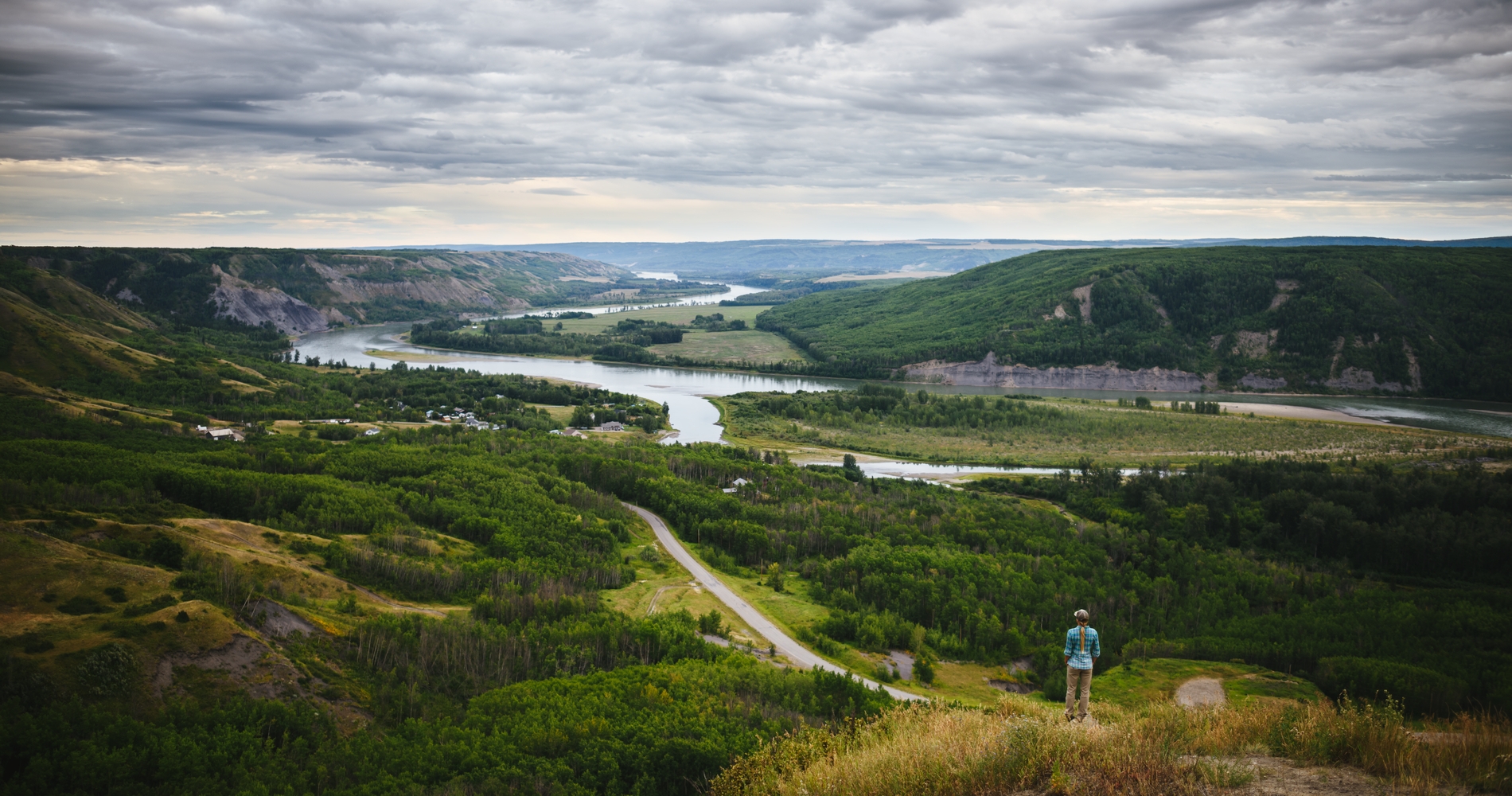 A person in a blue jacket stand on a hill overlooking a fertile river valley