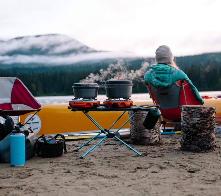 Campers cooking and camping along the banks in Bowron Lake Provincial Park. Two pots are bowling on a camping stove as the campers look out across the lake by their yellow canoe.