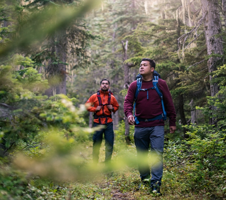 Two men hiking in Burnie-Shea Provincial Park. They are each wearing a backpack and different shades of red, long sleeve tops.