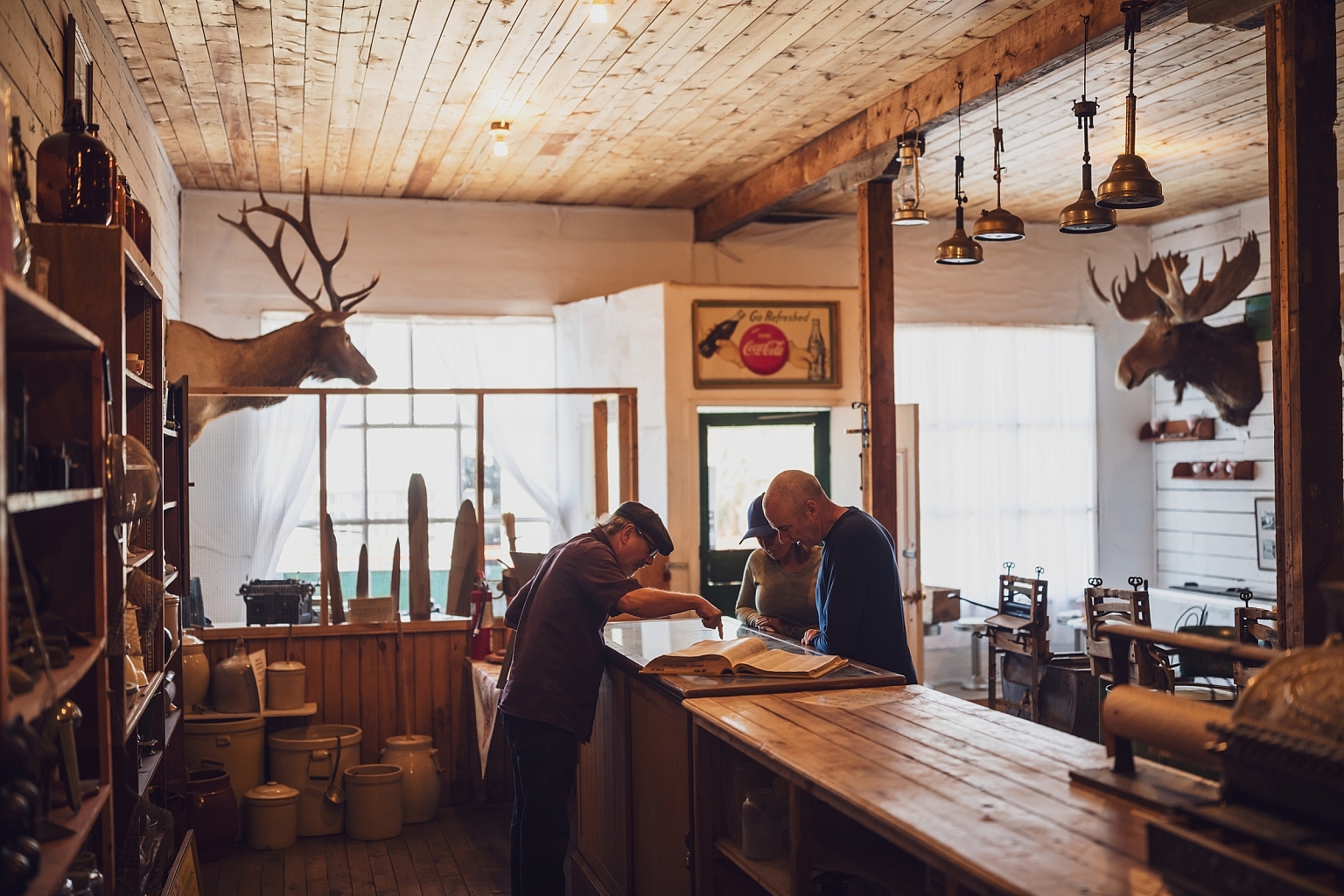 A room with a deer head on the wall, wooden ceiling, and an old time feel. Two visitors are interacting with a costumed staff person.