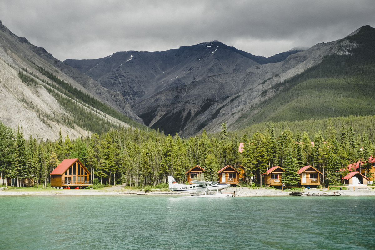 A sea plane sits on the surface of a lake near the shore, which is lined with wooden cabins. Mountains tower in the background.