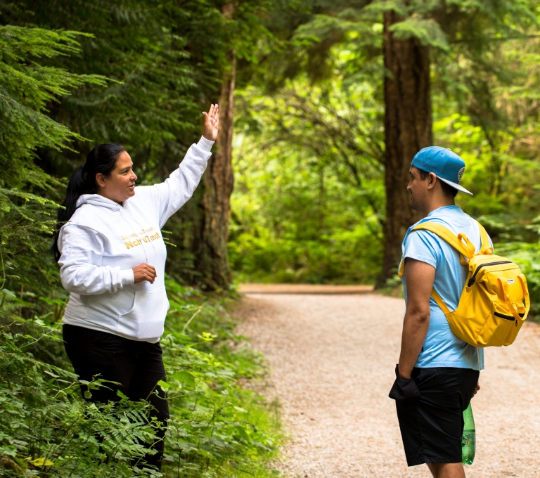 An Indigenous interpreter points to the trees to a tourist. They are standing on a path in Stanley Park.