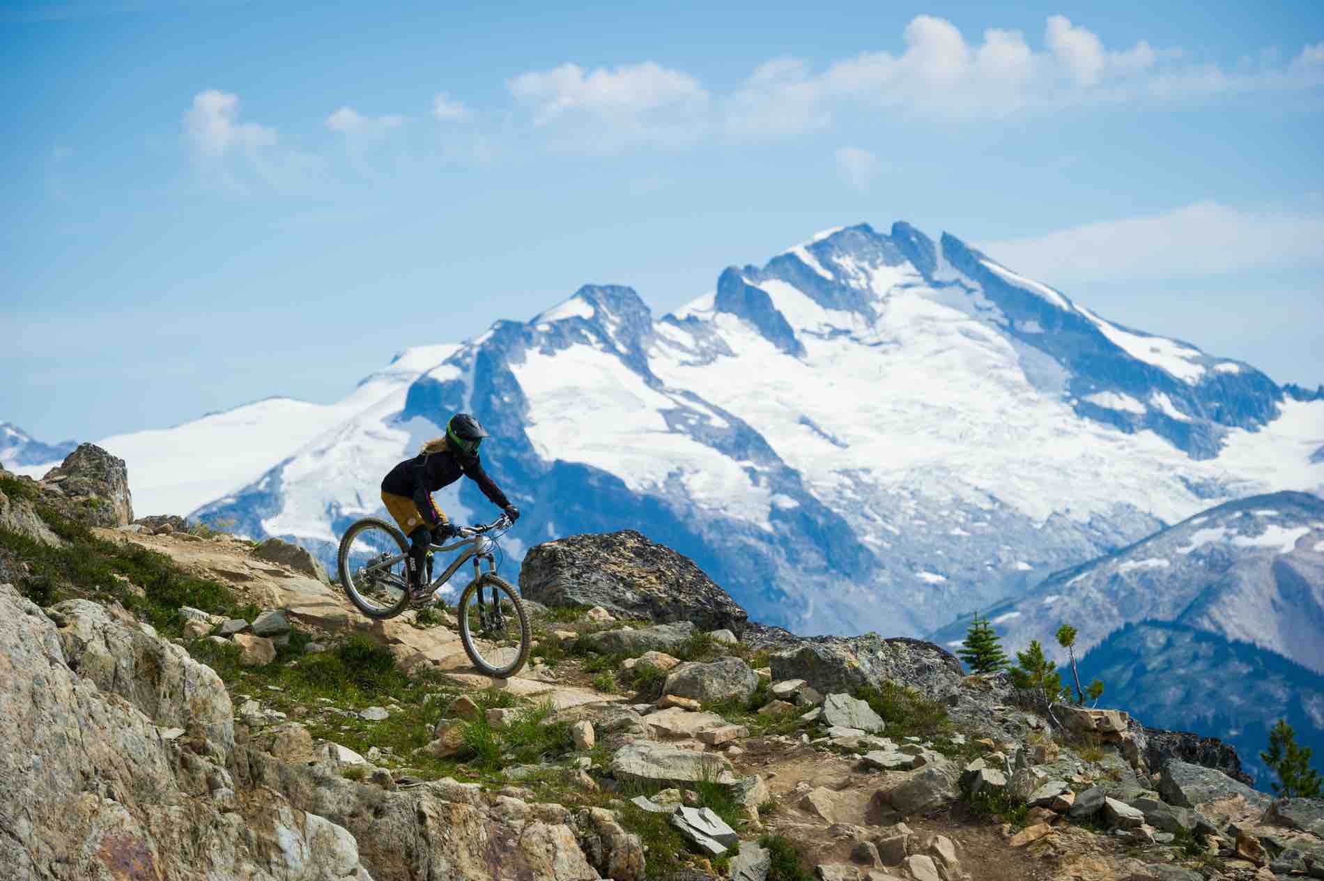 A person mountain bikes in the alpine down a rocky dirt path on the Whistler Mountain Bike Park. A snow covered mountain is in the background.