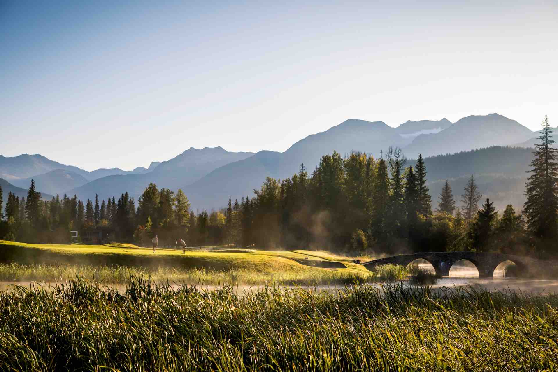 Two golfers in the distance on the green. Whistler and Blackcomb mountains are seen behind the golf course.