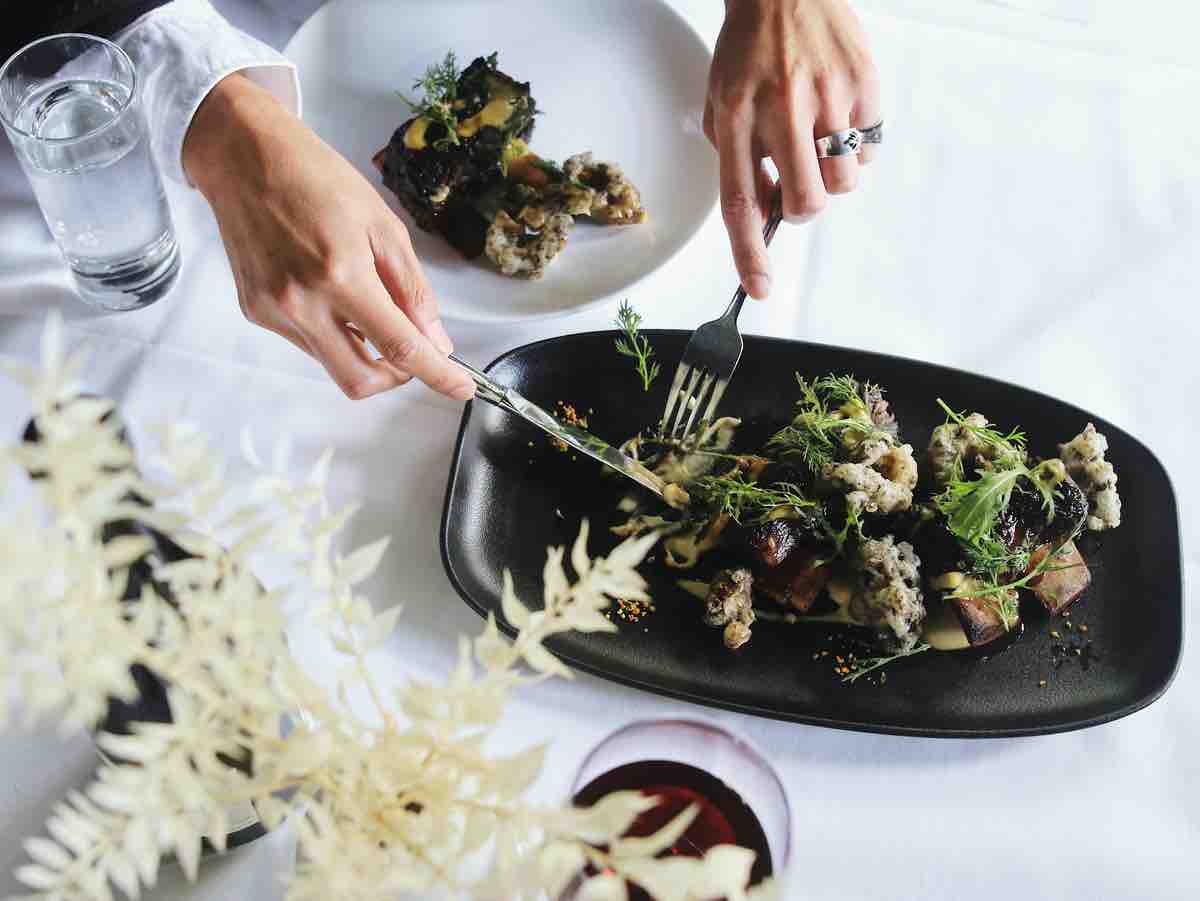 A person uses a knife and fork to cut a piece of cauliflower and salad from a shared plate, only their hands are seen. The table has a white linen table cloth. There is a glass of red wine and a white plant on then table.