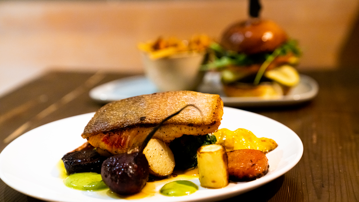 Two plates of food on a table. One is plated with salmon and vegetables, the other has a burger and fries.