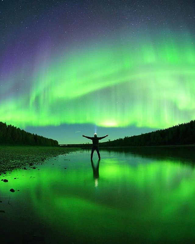A person stands in a shallow river and looks up at the Northern Lights in the night sky