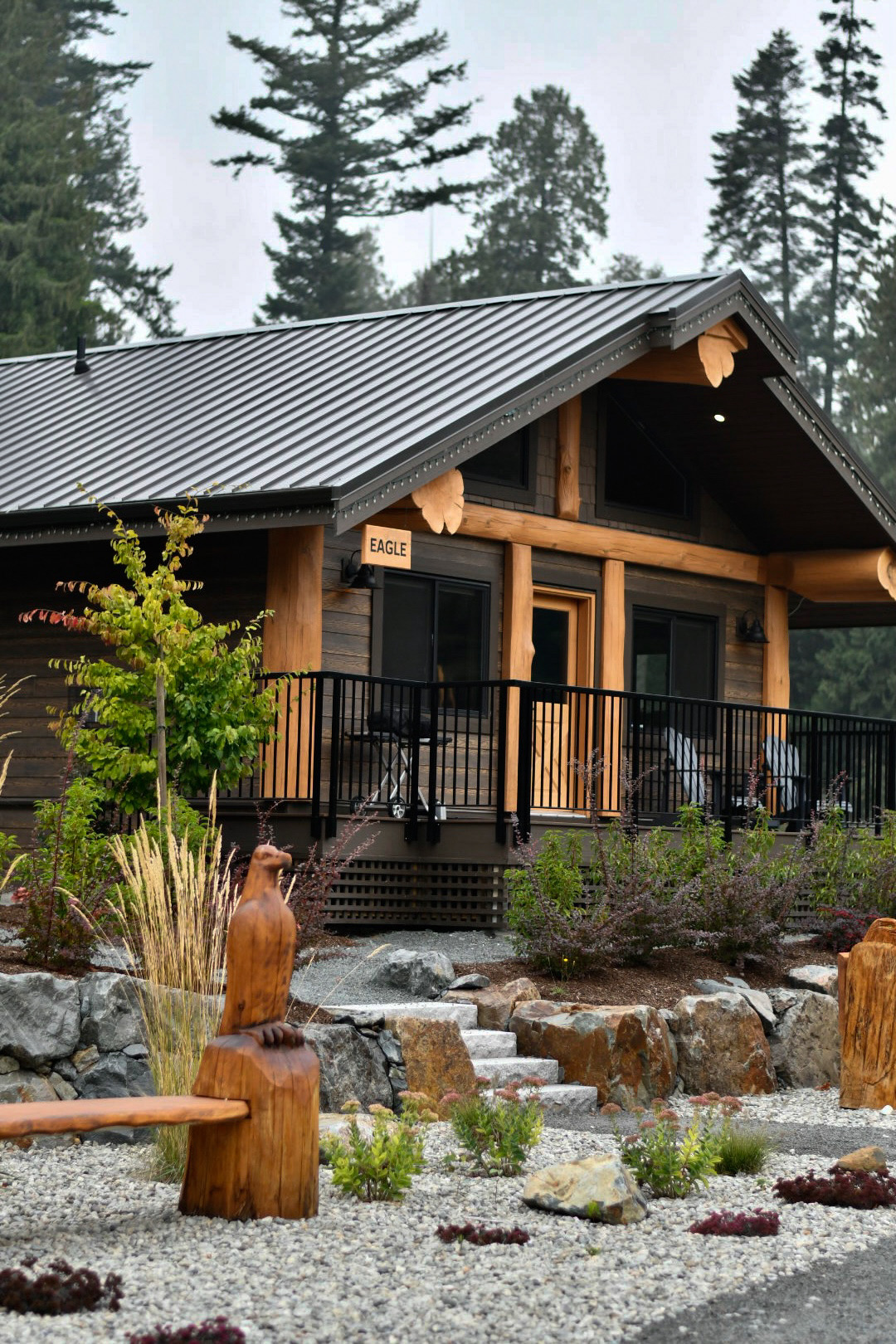 View of a wood building with a slanted metal roof, a black railing along the porch, with a sparse rock garden in front