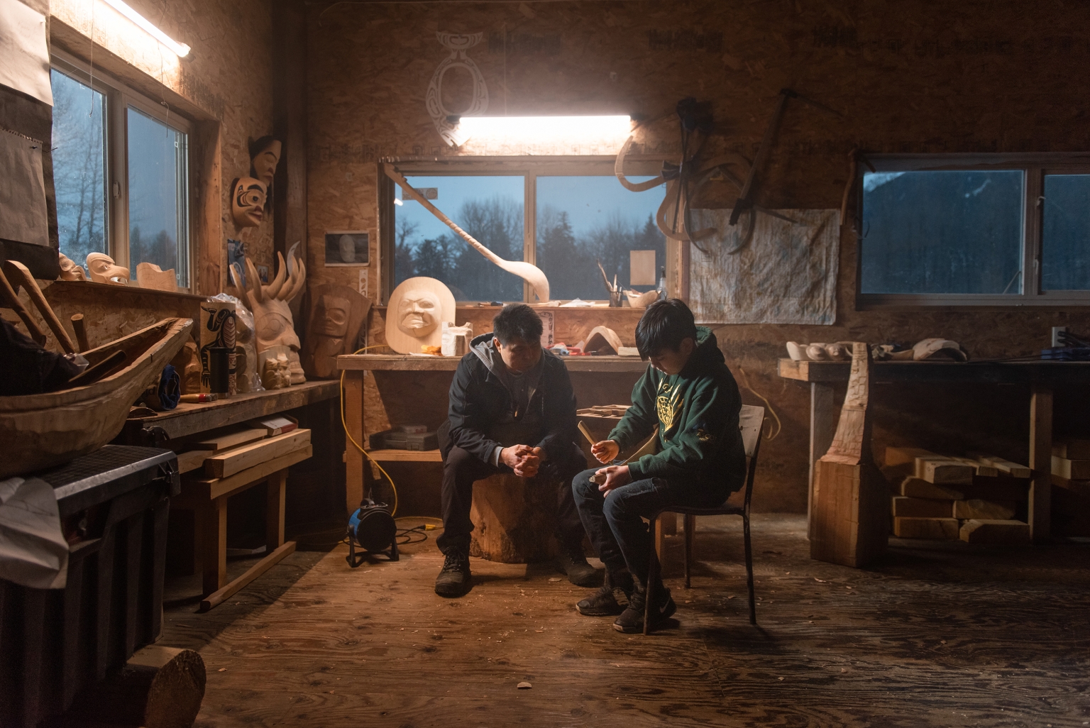 An Indigenous carver sits in a workshop next to a young boy who looks to be learning to carve