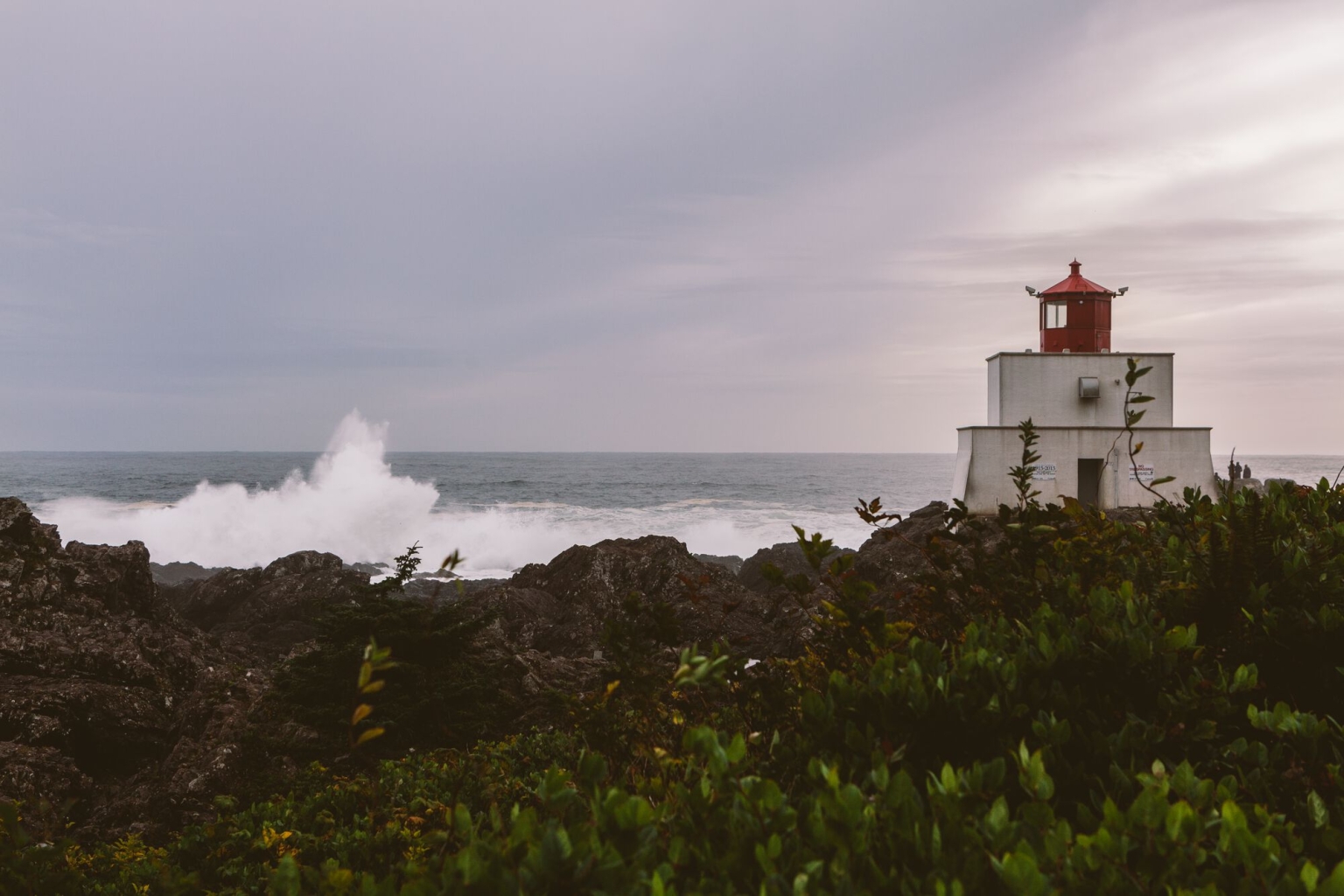 A short white lighthouse with a red top sits on the right side of the image o a rocky shorline with waves breaking in the background.