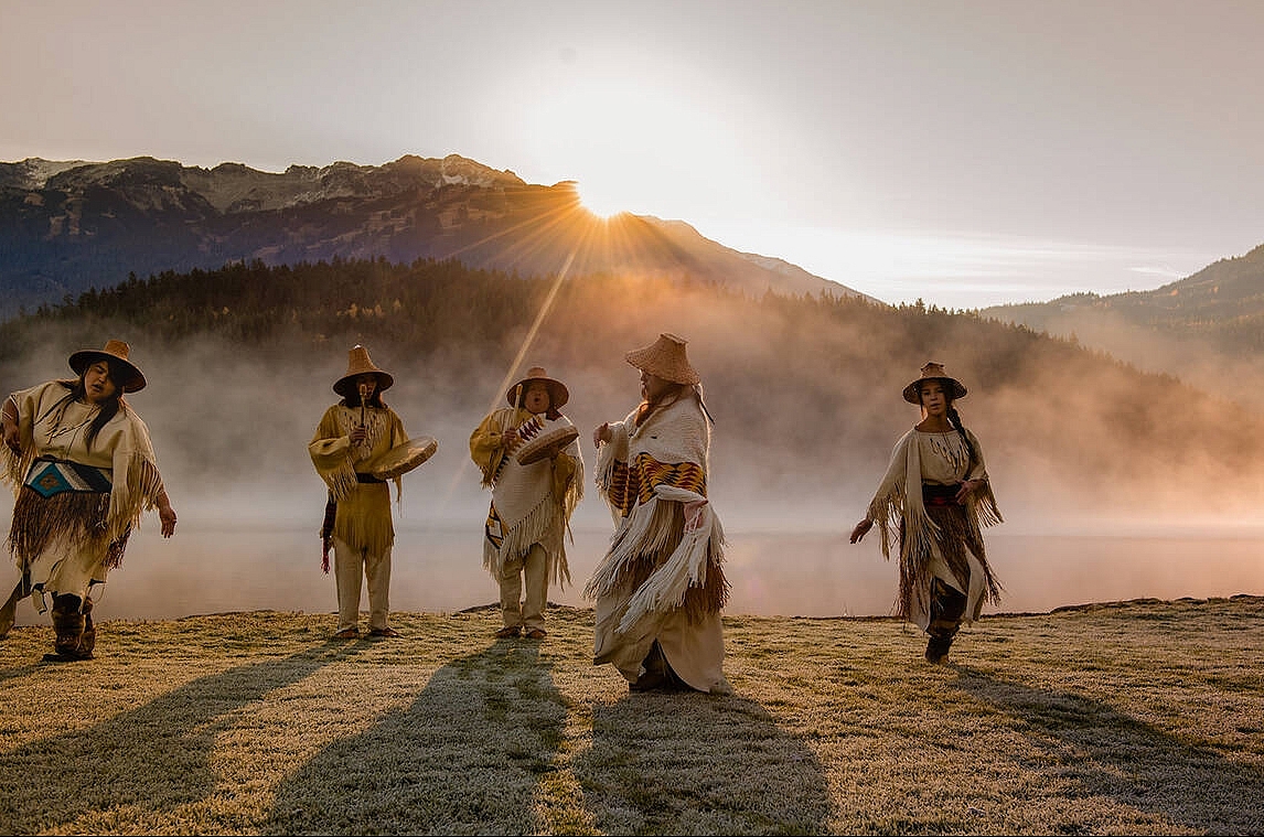 Five Indigenous people in traditional attire dance and play drums in a field with mist rising and the sun peeking over the mountains in the distance