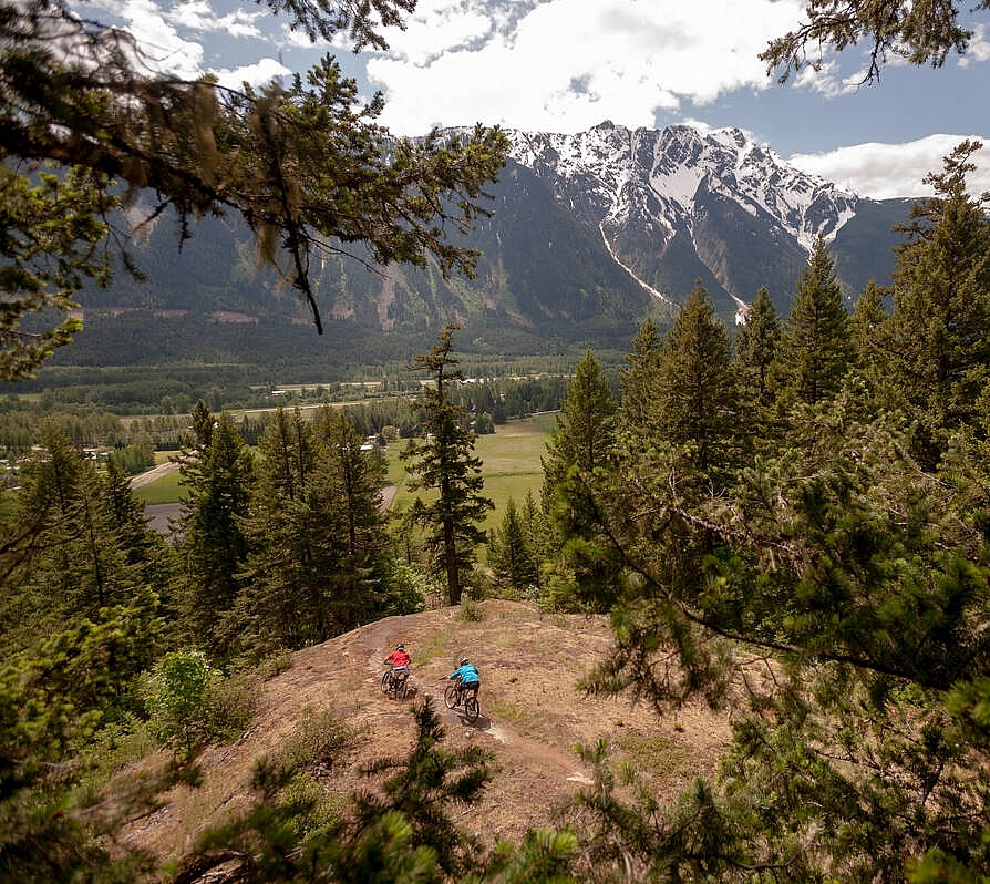 Two mountain bikers ride along a dirt trail overlooking a green valley with mountains in the background